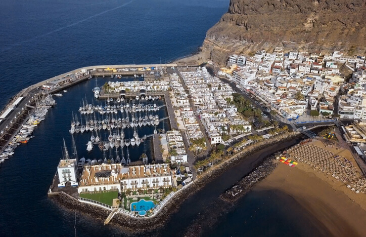 Flygfoto över Puerto de Mogan stad med flera vita hus och en hamn med massa segelbåtar som är skyddad med en vall från havet.